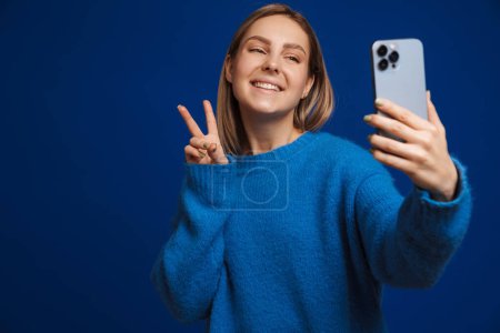 Téléchargez les photos : Jeune belle fille souriante prenant selfie montrant geste de victoire debout sur fond bleu isolé - en image libre de droit