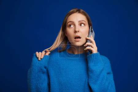 Foto de Joven chica atractiva juega con su cabello con la boca abierta mientras habla teléfono sobre fondo azul aislado - Imagen libre de derechos