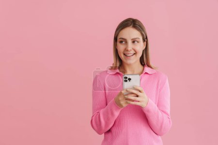 Foto de Joven chica guapa en suéter rosa sosteniendo el teléfono y mirando hacia la izquierda de pie sobre fondo aislado rosa - Imagen libre de derechos