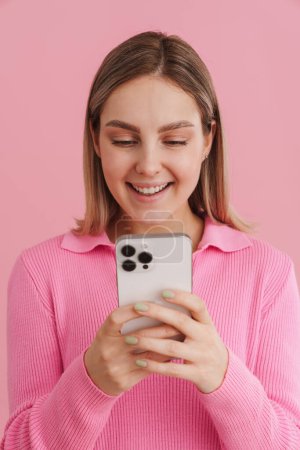 Foto de Joven chica guapa en suéter rosa sosteniendo el teléfono y mirándolo de pie sobre fondo aislado rosa - Imagen libre de derechos