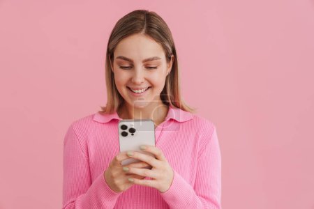 Téléchargez les photos : Jeune belle fille en pull rose tenant le téléphone et la regardant debout sur fond isolé rose - en image libre de droit