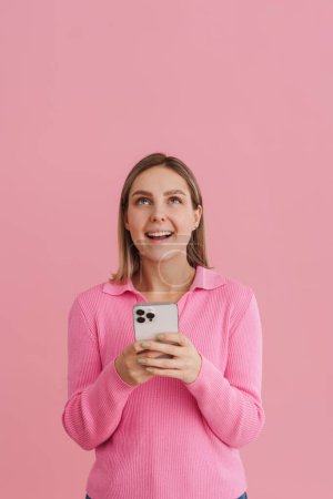 Téléchargez les photos : Jeune belle fille en pull rose tenant le téléphone et regardant vers le haut avec la bouche ouverte debout sur fond isolé rose - en image libre de droit