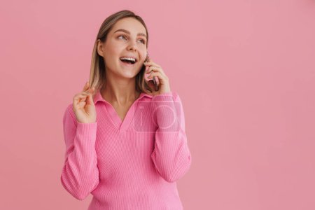 Foto de Joven chica atractiva juega con su cabello con la boca abierta mientras habla teléfono sobre fondo aislado rosa - Imagen libre de derechos