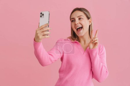 Foto de Joven hermosa mujer blanca sonriendo y tomando foto selfie en el teléfono celular de pie aislado sobre fondo rosa - Imagen libre de derechos