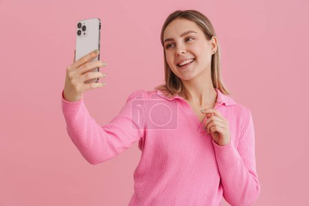 Téléchargez les photos : Jeune belle fille heureuse souriante prenant selfie en pull rose sur fond bleu isolé - en image libre de droit