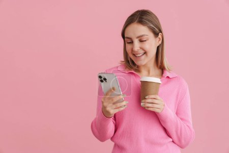 Téléchargez les photos : Jeune belle femme blanche tenant une tasse de café et utilisant un téléphone portable debout isolé sur fond rose - en image libre de droit