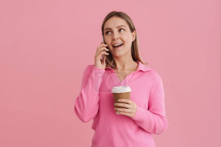 Téléchargez les photos : Jeune belle femme blanche tenant tasse de café et parlant sur téléphone portable debout isolé sur fond rose - en image libre de droit