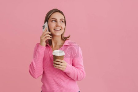 Téléchargez les photos : Jeune belle femme blanche tenant tasse de café et parlant sur téléphone portable debout isolé sur fond rose - en image libre de droit