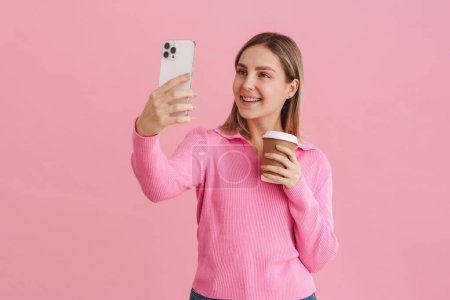 Photo for Young beautiful white woman holding coffee cup and taking selfie photo on cellphone standing isolated over pink background - Royalty Free Image