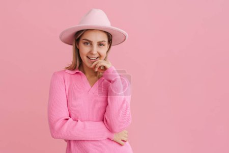 Foto de Joven hermosa dama en sombrero rosa y suéter tocando su barbilla y mirando en la cámara de pie sobre el fondo rosa aislado - Imagen libre de derechos