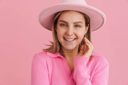 Foto de Joven hermosa dama en sombrero rosa y suéter tocando su mejilla y mirando en la cámara de pie sobre el fondo rosa aislado - Imagen libre de derechos