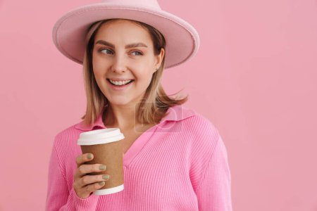 Foto de Joven hermosa dama en sombrero rosa y suéter con café mirando hacia la derecha y de pie sobre fondo rosa aislado - Imagen libre de derechos
