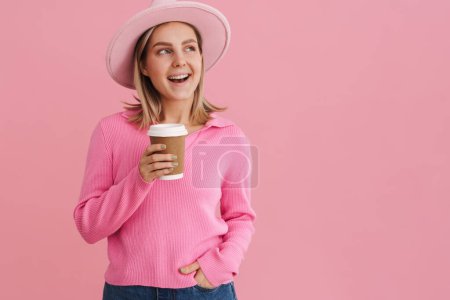 Photo for Young beautiful lady in pink hat and sweater with coffee looking rightward and standing over isolated pink background - Royalty Free Image