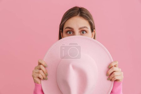 Téléchargez les photos : Jeune fille attrayante se cachant derrière un chapeau rose sur fond isolé rose - en image libre de droit
