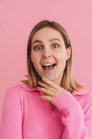 Foto de Joven chica sorprendida tocando su barbilla con la boca abierta mirando en la cámara y de pie sobre el fondo rosa aislado - Imagen libre de derechos
