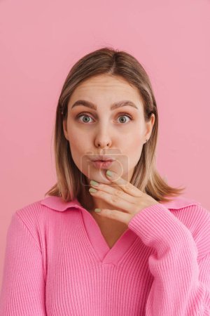 Foto de Joven chica sorprendida tocándose la barbilla con labios fruncidos mirando a la cámara y de pie sobre un fondo rosa aislado - Imagen libre de derechos