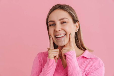 Foto de Joven linda chica sonriente tocando sus dedos índice en sus mejillas sobre el fondo rosa aislado - Imagen libre de derechos