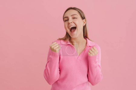 Foto de Joven mujer blanca hermosa usando camisa gritando como ganador y apretando los puños de pie aislado sobre fondo rosa - Imagen libre de derechos