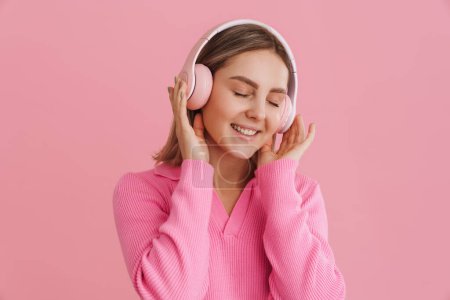 Foto de Joven chica sonriente beutiful disfrutando de la música con los ojos cerrados en auriculares de color rosa sobre fondo rosa aislado - Imagen libre de derechos