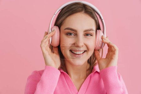 Foto de Joven chica sonriente beutiful disfrutando de la música en auriculares de color rosa mirando en la cámara sobre el fondo rosa aislado - Imagen libre de derechos