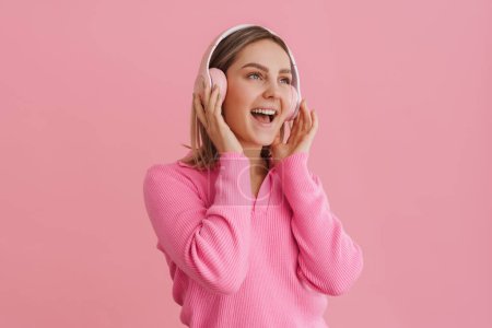 Foto de Joven chica entusiasta con la boca abierta en auriculares de color rosa de pie sobre el fondo rosa aislado - Imagen libre de derechos