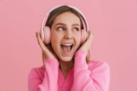 Foto de Joven chica entusiasta con la boca abierta en auriculares de color rosa de pie sobre el fondo rosa aislado - Imagen libre de derechos