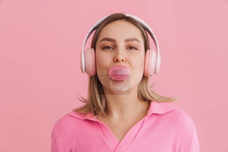 Foto de Joven linda chica en auriculares rosados soplando goma de mascar sobre fondo rosa aislado - Imagen libre de derechos