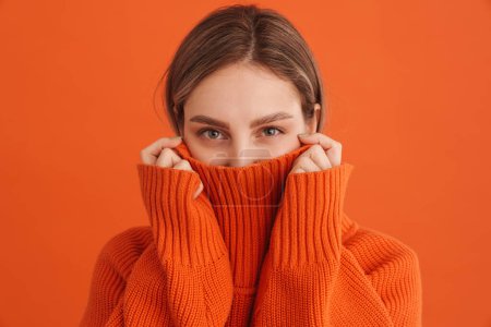 Foto de Joven linda chica escondida en suéteres cuello sobre fondo naranja aislado - Imagen libre de derechos
