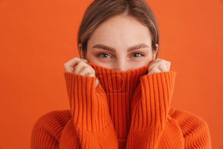 Foto de Joven linda chica escondida en suéteres cuello sobre fondo naranja aislado - Imagen libre de derechos