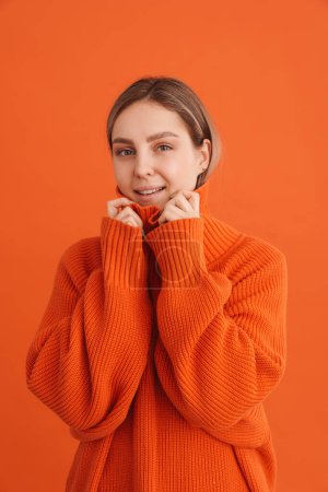 Foto de Joven linda chica sonriente sosteniendo suéteres cuello sobre fondo naranja aislado - Imagen libre de derechos