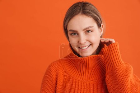 Foto de Joven hermosa chica en suéter naranja tocando su mejilla y mirando en la cámara de pie sobre el fondo naranja aislado - Imagen libre de derechos