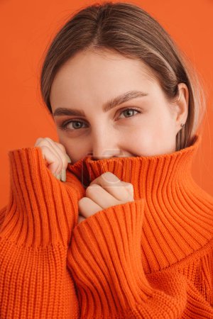 Foto de Retrato de una joven linda chica escondida en suéteres cuello sobre fondo naranja aislado - Imagen libre de derechos