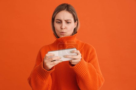 Foto de Joven linda chica enfocada jugando juego móvil en su teléfono de pie sobre fondo naranja aislado - Imagen libre de derechos