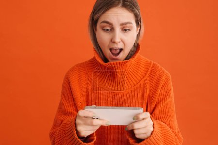 Téléchargez les photos : Jeune belle fille enthousiaste jouant jeu mobile sur son téléphone debout sur fond orange isolé - en image libre de droit