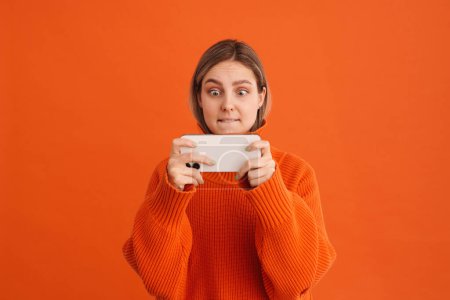 Téléchargez les photos : Jeune belle fille tendue jouant jeu mobile sur son téléphone debout sur fond orange isolé - en image libre de droit