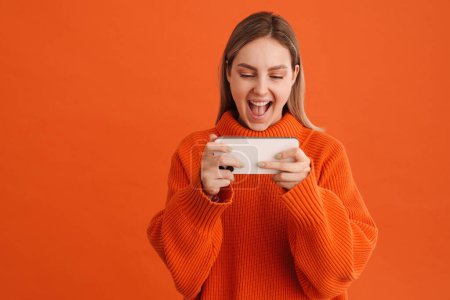 Foto de Joven hermosa chica sonriente feliz jugando juego móvil en su teléfono de pie sobre fondo naranja aislado - Imagen libre de derechos