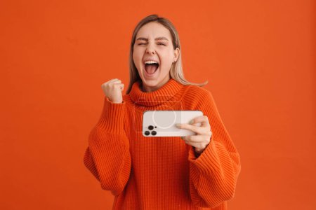 Photo for Young beautiful enthusiastic girl celebrating victory in mobile game on her phone standing over isolated orange background - Royalty Free Image