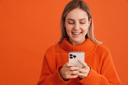 Foto de Joven chica guapo en suéter naranja sosteniendo el teléfono y mirando de pie sobre fondo aislado naranja - Imagen libre de derechos