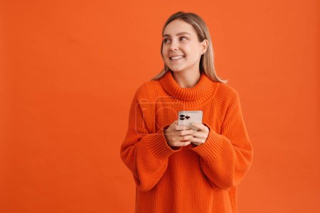 Foto de Joven chica guapa en suéter naranja sosteniendo el teléfono y mirando hacia la izquierda de pie sobre fondo aislado naranja - Imagen libre de derechos