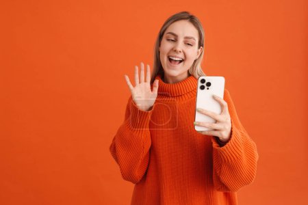 Foto de Joven hermosa chica tomando selfie o haciendo videocall saludando a la cámara sobre fondo naranja aislado - Imagen libre de derechos