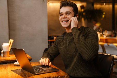 Téléchargez les photos : Jeune homme blanc portant pull souriant tout en utilisant ordinateur portable et téléphone portable dans le café - en image libre de droit