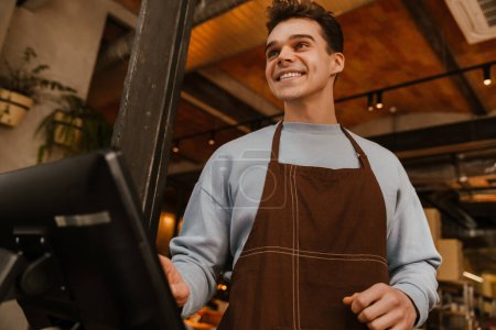 Téléchargez les photos : Jeune homme serveur blanc portant tablier debout devant l'ordinateur et enregistrant le paiement dans le café - en image libre de droit