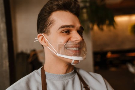 Foto de Joven camarero blanco con delantal y mascarilla sonriendo mientras trabaja en la cafetería - Imagen libre de derechos