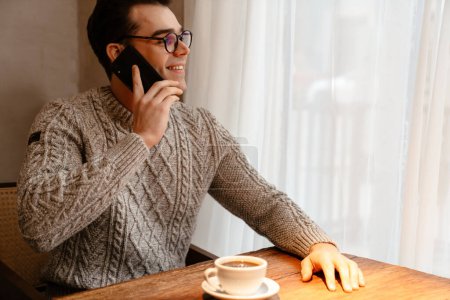 Téléchargez les photos : Jeune homme blanc portant des lunettes souriant tout en buvant du café et en utilisant un téléphone portable dans le café - en image libre de droit