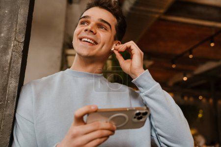 Téléchargez les photos : Jeune homme blanc heureux portant des écouteurs souriant et utilisant un téléphone portable dans le café - en image libre de droit