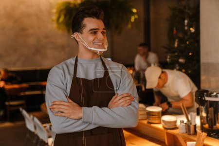 Photo for Young white waiter man wearing apron and face mask smiling while working in cafe - Royalty Free Image