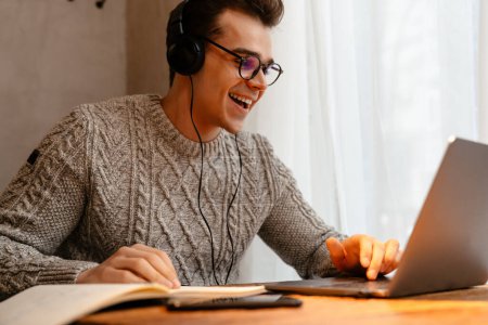 Téléchargez les photos : Jeune homme blanc portant des lunettes souriant tout en utilisant un ordinateur portable et un casque dans le café - en image libre de droit