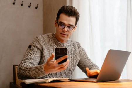 Téléchargez les photos : Jeune homme blanc portant des lunettes de vue travaillant sur un ordinateur portable et prenant des notes tout en étant assis dans un café - en image libre de droit