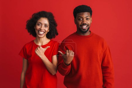 Téléchargez les photos : Noir heureux homme et femme souriant et gesticulant à l'autre isolé sur fond rouge - en image libre de droit