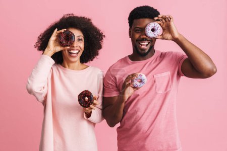 Téléchargez les photos : Noir homme et femme souriant et s'amusant avec des beignets isolés sur fond rose - en image libre de droit